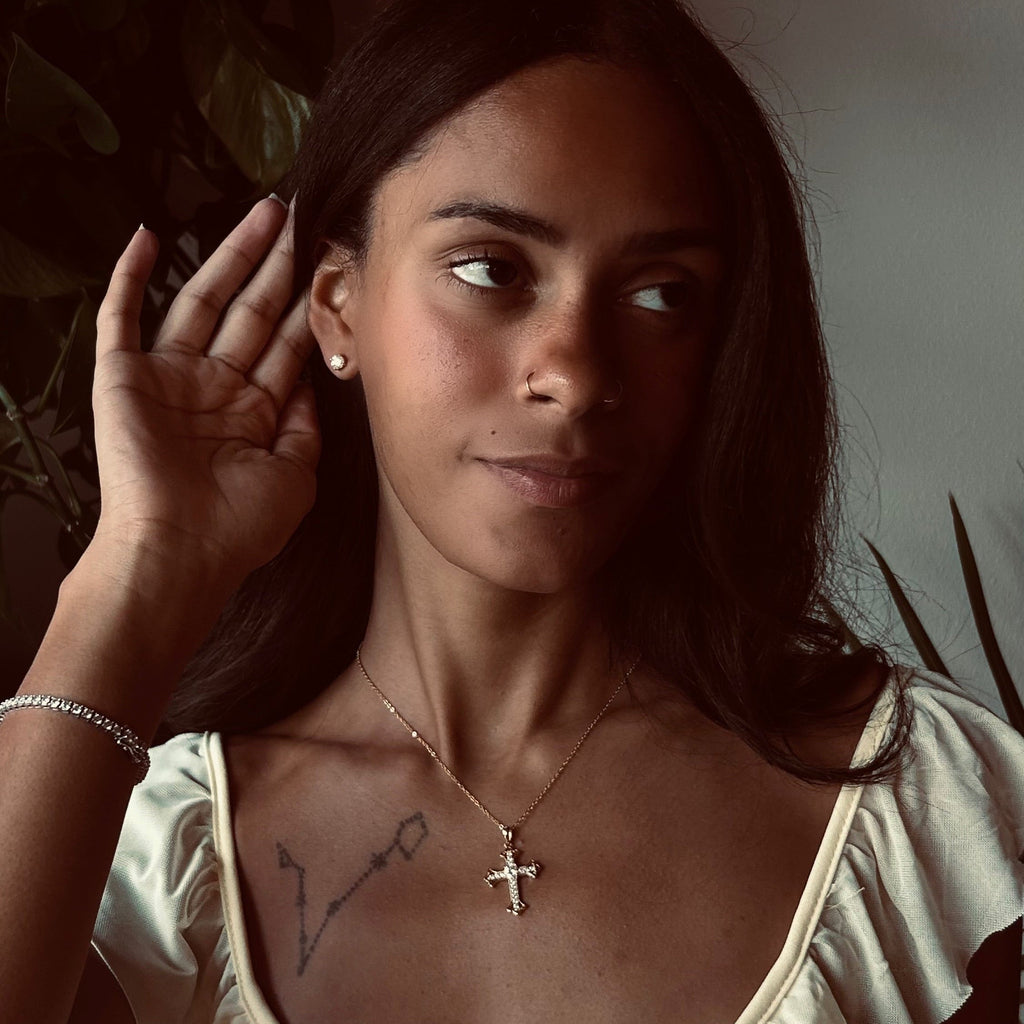 A woman wearing a delicate silver cross pendant necklace on a fine chain, styled with a butter yellow dress. She is posed with warm, dim lighting, looking off-camera with one hand near her ear to expose her earring.